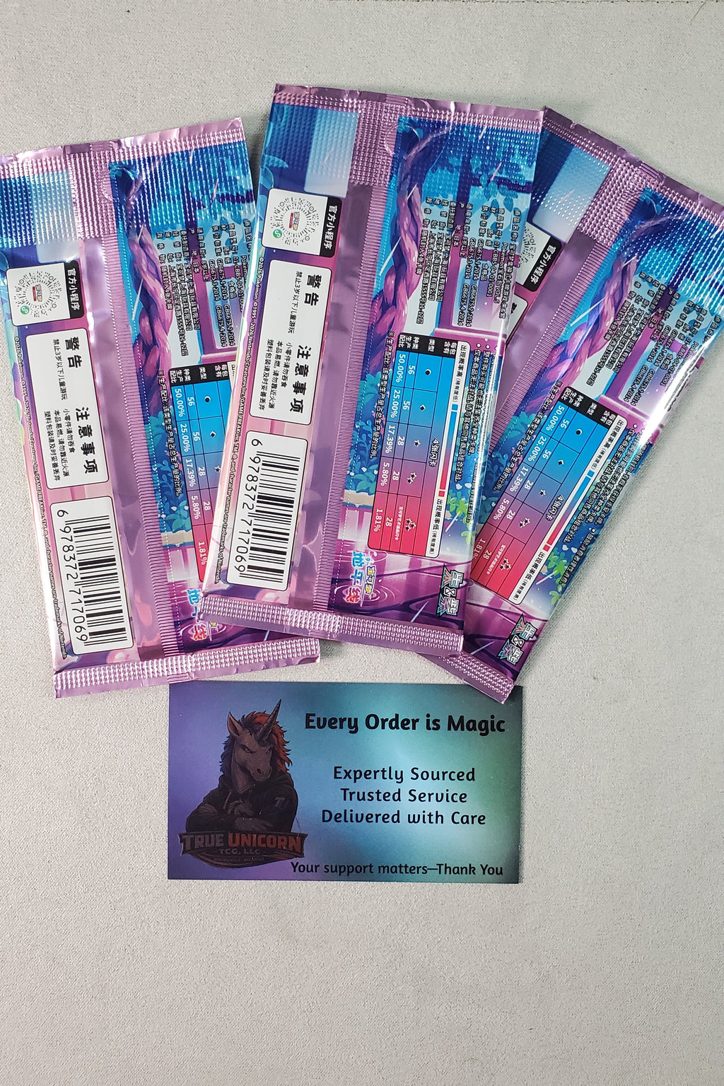 Three Chinese Pokémon booster packs on a light gray surface, with a small printed card below them that reads 'Every Order is Magic' and shows a True Unicorn Logo illustration. The packs have purple, blue, and pink packaging with holographic graphics.