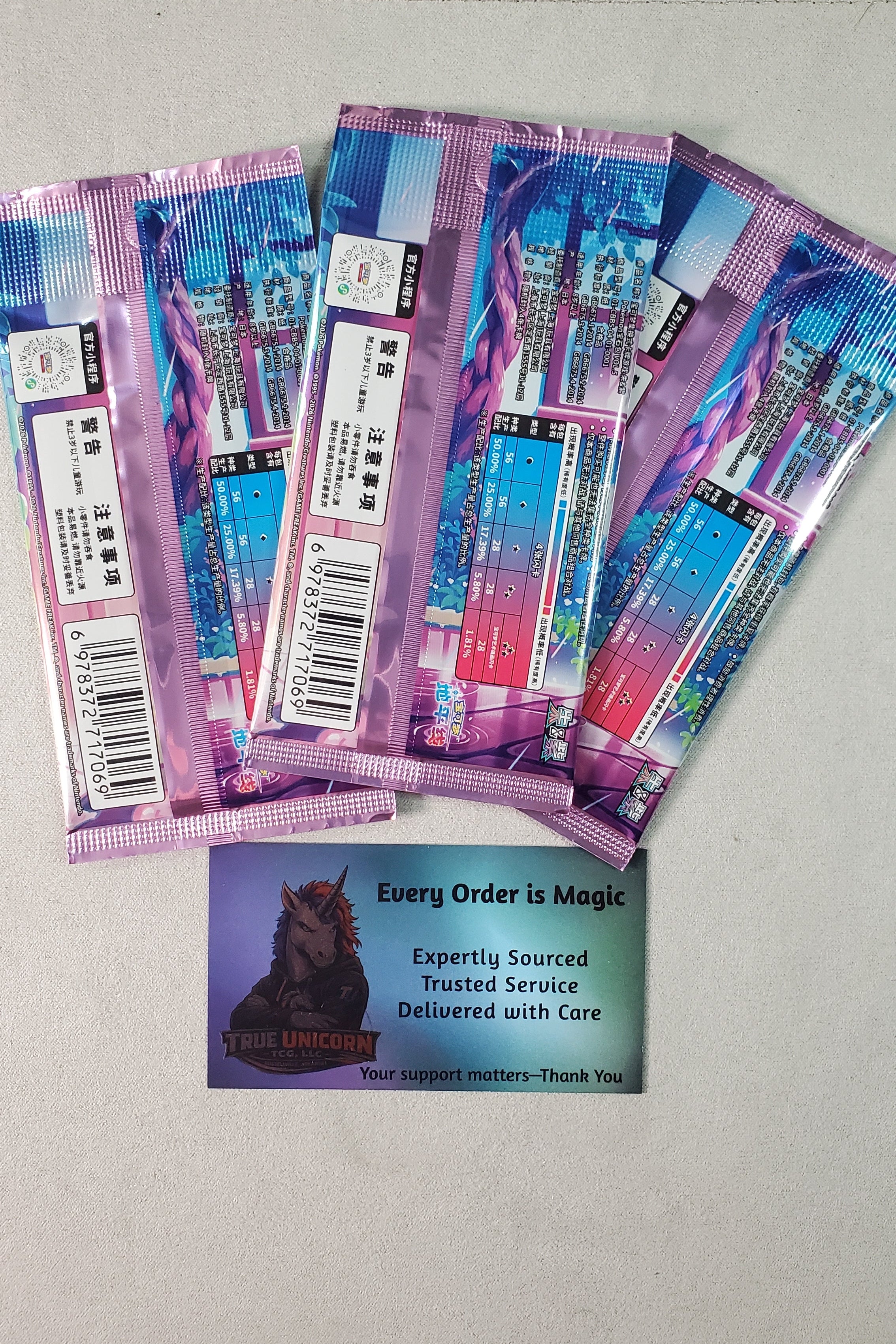 Three Chinese Pokémon booster packs on a light gray surface, with a small printed card below them that reads 'Every Order is Magic' and shows a True Unicorn Logo illustration. The packs have purple, blue, and pink packaging with holographic graphics.