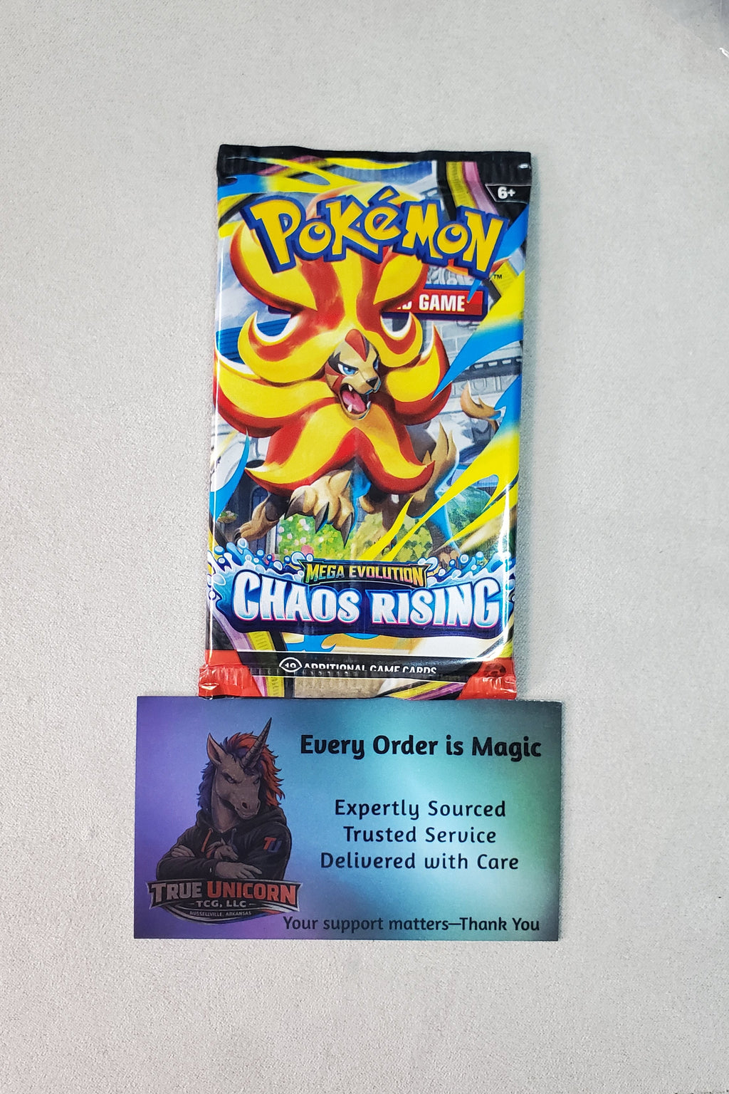 A Pokémon TCG booster pack for 'Chaos Rising' shown above a custom 'True Unicorn' support card on a light gray desk surface.
