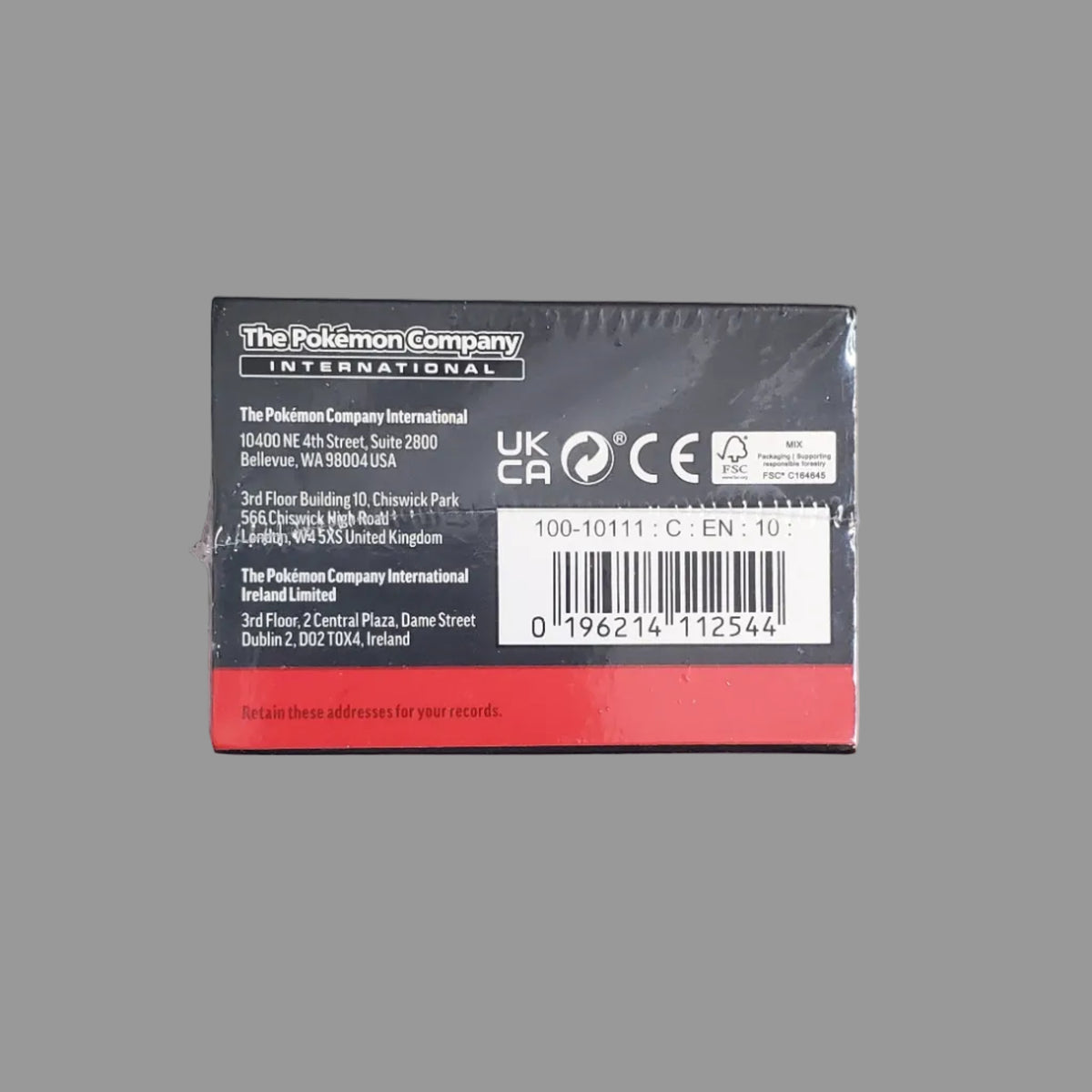 A small rectangular product box photographed from above on a light gray surface. The box is wrapped in clear plastic and has a black top panel with white text, a barcode, and a red band along the bottom edge.
