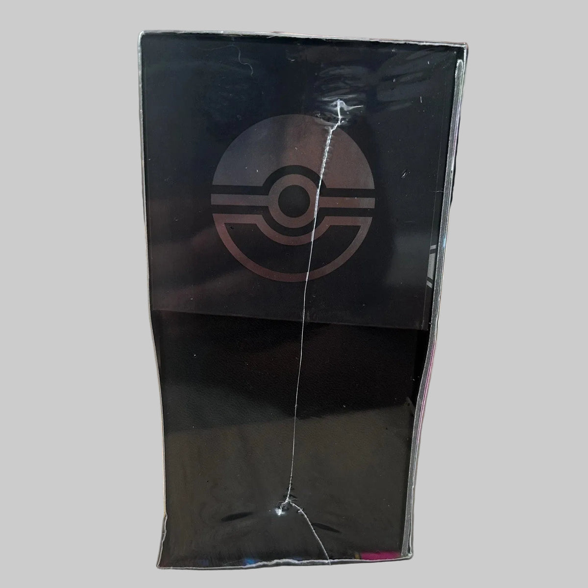 A black rectangular product box standing upright on a light wood surface, featuring a large Pokémon ball logo on the front. The box is wrapped in clear plastic with visible scuffs and a vertical crack, and the background shows a blurred indoor setting.