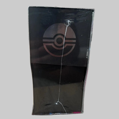 A black rectangular product box standing upright on a light wood surface, featuring a large Pokémon ball logo on the front. The box is wrapped in clear plastic with visible scuffs and a vertical crack, and the background shows a blurred indoor setting.