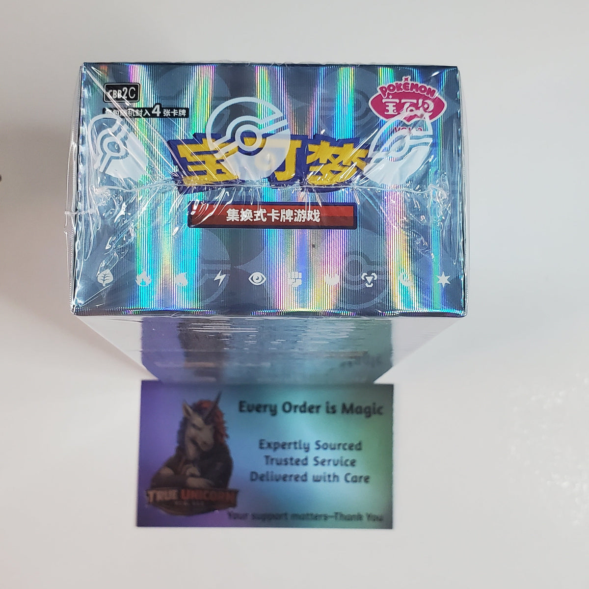 A sealed holographic blue box of Chinese collectible trading cards, with a purple insert card partially visible below it against a plain white wall.