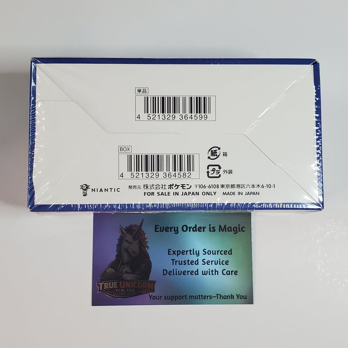 A sealed white product box with blue trim and printed barcodes, photographed on a plain light background. A small printed card with a horse illustration and the text 'Every Order is Magic' is placed in front of the box.