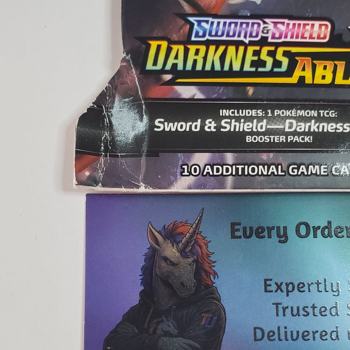 A Pokémon TCG booster pack for Sword & Shield: Darkness Ablaze shown above a Blue Unicorn TCG card on a plain white background.