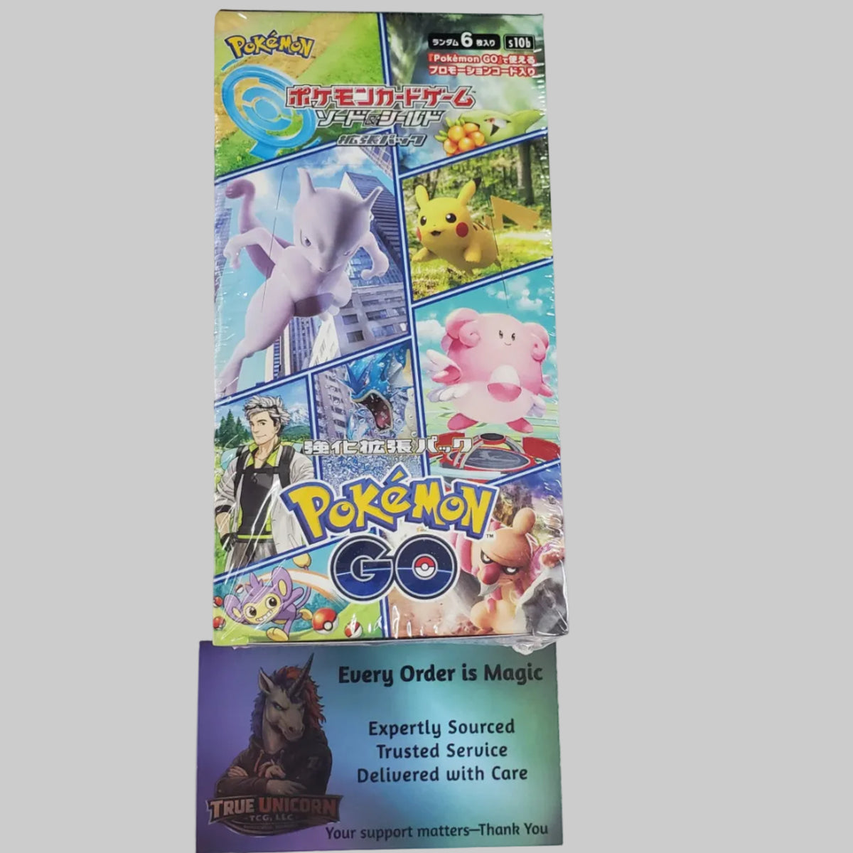 A Pokémon Go booster box standing upright on a white surface, with a smaller purple Pokémon card pack placed underneath it. The box features colorful artwork of popular Pokémon characters and the Pokémon Go logo.