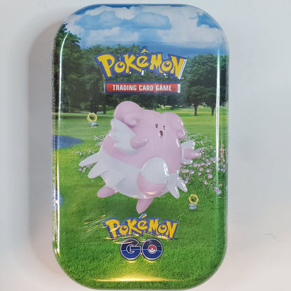 A rectangular Pokémon Trading Card Game tin featuring a grassy outdoor scene with a pink character in the center, photographed on a plain light background.